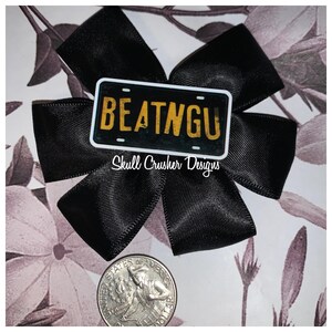 May include: A black satin bow with a license plate design in the center that reads "BEATNGU" in yellow lettering. The text "Skull Crusher Designs" is below the license plate. A quarter is in the foreground.