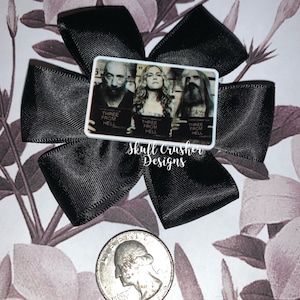 May include: A black bow with a white square featuring a movie poster for "Three From Hell" with three people in mugshots. The text "Skull Crusher Designs" is written below the square.