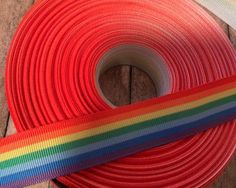 May include: A roll of red ribbon with a rainbow ribbon on top. The rainbow ribbon has stripes of red, orange, yellow, green, blue, indigo, and violet.