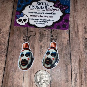 May include: A pair of dangling earrings featuring a clown face with a red and white top hat. The earrings are silver-toned and hang from a hook. A business card with the text "SKULL CRUSHER designs" is in the background.