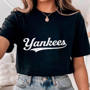 May include: Black t-shirt with the word "Yankees" in white, stylized script. The shirt is a classic crew neck style. The text has a distressed, vintage look.