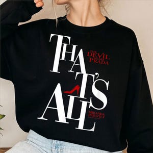 Puede incluir: Sudadera negra con el texto "THAT'S ALL" en letras blancas. Las palabras "THE DEVIL WEARS PRADA" son rojas, con un gráfico de zapato de tacón alto rojo. La cita "-MIRANDA PRIESTLY" es roja.