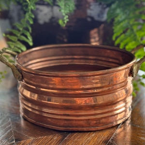 May include: A copper-colored, round planter with two brass handles. The planter has horizontal ridges around the body. The background includes a dark brown pot with green plants and a wooden surface.