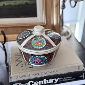 May include: A decorative ceramic jar with a lid, featuring a brown base and lid with white stripes and floral designs in shades of blue, red, and yellow. The jar is placed on top of a stack of books.