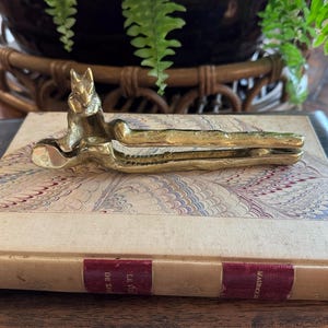 May include: A brass nutcracker shaped like a fox, featuring a metallic gold finish and a textured, elongated handle design. The tool is constructed with a hinged mechanism for cracking nuts, displaying a detailed fox head at the top end.