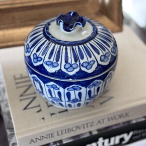May include: A small, round ceramic jar with a lid, featuring a blue and white floral pattern. The jar is sitting on top of a stack of books. The lid has a small, ruffled handle. The jar's design includes vertical lines and floral motifs.