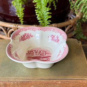May include: A cream-colored ceramic bowl with a scalloped edge and a pink transferware pattern. The interior features a detailed landscape illustration of a castle, while the rim is decorated with floral motifs and intricate scrollwork.