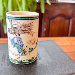 May include: A cylindrical ceramic cup with a cream-colored crackle glaze finish. The exterior features a hand-painted illustration of a person in blue and white clothing carrying a large bundle of sticks, accented by green, pink, and orange details.