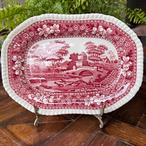 May include: An oval ceramic platter featuring a deep red and white transferware pattern. The central scene depicts a stone bridge, a castle, and trees. The wide, scalloped rim is decorated with a floral border. The platter rests on a metal stand.