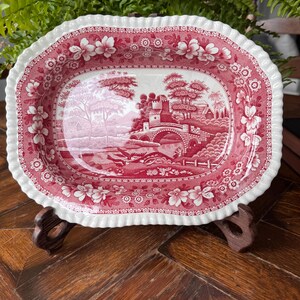 May include: An oval ceramic serving dish with a scalloped edge, featuring a red and white transferware pattern. The central design depicts a landscape with a stone bridge, a castle, and trees, while the border is decorated with floral motifs.