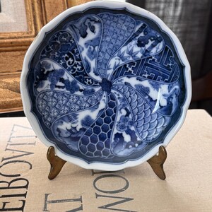 May include: A small, round, scalloped-edge ceramic dish featuring a blue and white pattern. The design consists of multiple radiating segments, each displaying distinct geometric, floral, and landscape motifs in a traditional cobalt blue glaze.