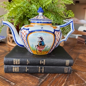 May include: A white ceramic teapot with a lid, featuring hand-painted blue, orange, and yellow floral and geometric patterns. The central panel depicts a figure in a hat and colorful clothing walking with a cane. The teapot has a curved spout and handle.