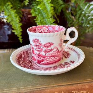 May include: A white porcelain teacup and saucer set featuring a detailed red transferware pattern of a landscape with trees, a small house, and a bridge. The rim of the cup and the edge of the saucer have a scalloped, embossed texture design.