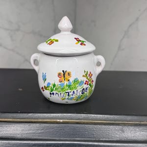 May include: A white ceramic mustard pot with a matching lid, featuring hand-painted colorful floral motifs, a central orange butterfly, and the word MOUTARDE written in blue. The pot has two small side handles and a pointed lid finial.