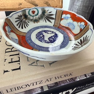 May include: A shallow, scalloped ceramic bowl featuring a central blue circular motif with a geometric border. The interior is decorated with orange, green, and brown painted patterns, including stylized pine tree branches and floral elements.