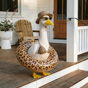 May include: A whimsical white goose statue wearing a leopard print bikini, sunglasses, and a matching hat. The goose is surrounded by a leopard print inflatable ring, ready for a day at the beach. The statue has yellow feet and a yellow beak.