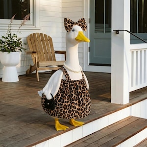 May include: A decorative goose figurine, dressed in a leopard print dress, bow, and a pearl necklace. The goose has a yellow beak and feet, and is holding a small black purse. The figurine is standing on a wooden porch.