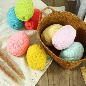 May include: A selection of colourful balls of yarn in shades of blue, green, red, pink, and yellow. Some are arranged on a white knitted surface, while others are in a woven basket. The yarn is suitable for knitting and crochet projects.
