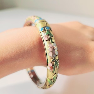 Puede incluir: Un brazalete dorado con detalles florales esmaltados. El brazalete presenta flores y hojas rosas, azules y verdes. Una pieza de joyería clásica.