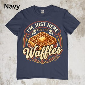 Puede incluir: Camiseta azul marino con un gr&aacute;fico de gofres. El gr&aacute;fico presenta una pila de gofres con mantequilla y el texto "I'M JUST HERE FOR THE Waffles". El dise&ntilde;o incluye colores naranja, amarillo y marr&oacute;n.