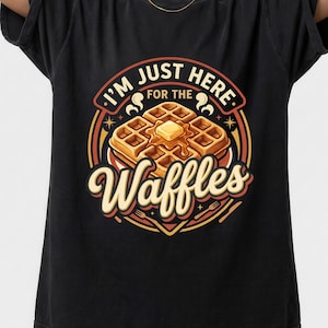 Puede incluir: Camiseta negra con un dise&ntilde;o gr&aacute;fico de un gofre con mantequilla y sirope, rodeado del texto "I'm just here for the Waffles". El dise&ntilde;o utiliza acentos naranjas, amarillos y rojos en un estilo retro.
