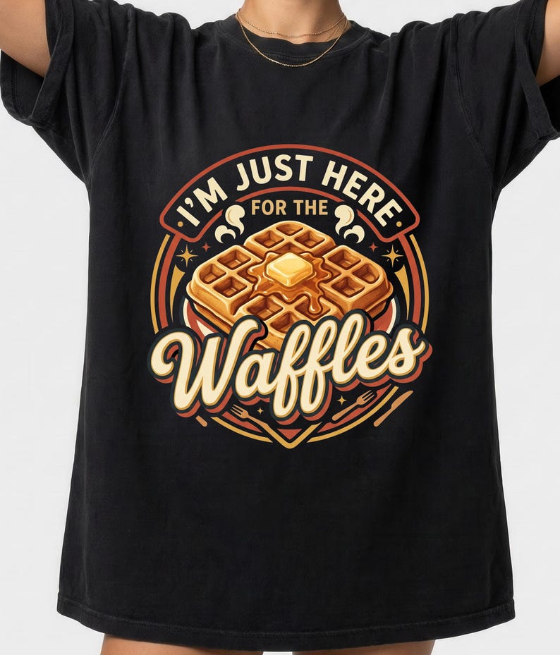 Puede incluir: Camiseta negra con un dise&ntilde;o gr&aacute;fico de un gofre con mantequilla y sirope, rodeado del texto "I'm just here for the Waffles". El dise&ntilde;o utiliza acentos naranjas, amarillos y rojos en un estilo retro.