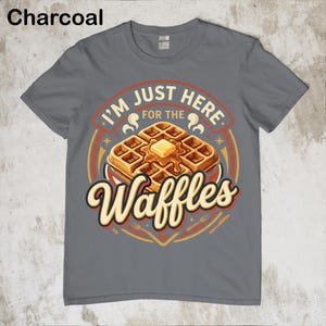 Puede incluir: Camiseta gris carb&oacute;n con un gr&aacute;fico de gofres. El gr&aacute;fico presenta una pila de gofres con mantequilla y el texto "I'm just here for the Waffles". El dise&ntilde;o es en tonos naranja, amarillo y marr&oacute;n.