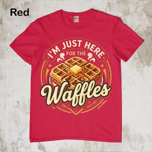 Puede incluir: Camiseta roja con un gr&aacute;fico de gofres. El gr&aacute;fico presenta una pila de gofres con mantequilla y el texto "I'M JUST HERE FOR THE Waffles". El dise&ntilde;o incluye un borde circular con estrellas e ilustraciones de tenedores y cuchillos.