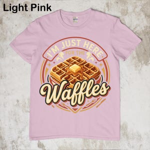 Puede incluir: Camiseta rosa claro con un dise&ntilde;o de gofre. El dise&ntilde;o dice "I'M JUST HERE FOR THE Waffles" en un c&iacute;rculo con un gofre con mantequilla. La camiseta es de cuello redondo.