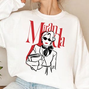 Puede incluir: Sudadera blanca con un gráfico rojo y negro de una mujer con gafas de sol, con el nombre "Miranda" en letras rojas. La ilustración muestra a una mujer sosteniendo libros, con una blazer y una pajarita.