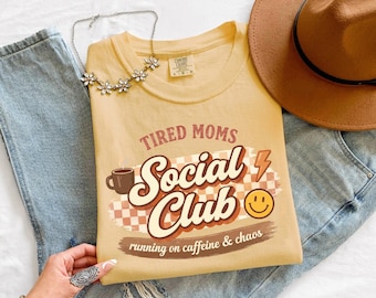 May include: A butter-colored t-shirt with the text "Tired Moms Social Club" in brown and white. The shirt features a coffee cup, lightning bolt, and smiley face graphic. The shirt is paired with blue jeans, a brown hat, and a floral necklace.