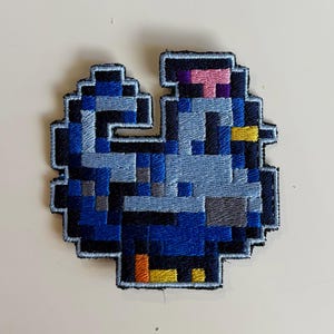 Può includere: Toppa ricamata con un disegno di uccello pixelato. La toppa utilizza sfumature di blu, grigio, nero, giallo e rosa. L'uccello ha un piccolo cappello rosa e un becco giallo. La toppa è probabilmente per abbigliamento o accessori.
