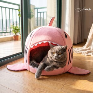 Puede incluir: Un gato gris descansa dentro de una cama para mascotas con forma de tiburón rosa. La cama tiene una boca abierta con dientes blancos y detalles de ojos negros. La cama tiene aletas y una cola. El gato está mirando a la cámara.