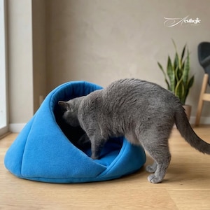 Pode incluir: Um gato cinzento explora uma cama para gatos azul brilhante. A cama tem um design macio e fechado, com uma forma arredondada. O gato é o foco principal, com a cama proporcionando um espaço aconchegante. O fundo inclui uma janela, uma planta e uma cadeira.