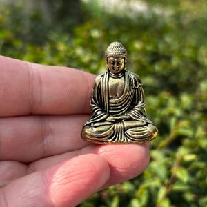 Könnte beinhalten: Kleine, detaillierte Buddha-Statue aus Messing in sitzender Meditationshaltung. Die Figur hat einen ruhigen Ausdruck, mit komplizierten Details in den Gewändern und Haaren. Die Statue wird in einer Hand gehalten.