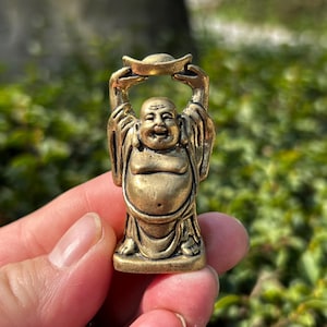 May include: A small, bronze-colored Buddha statue with a smiling face, holding a gold ingot above its head. The figure has a rounded belly and is wearing draped robes. The statue is a symbol of wealth and good fortune.