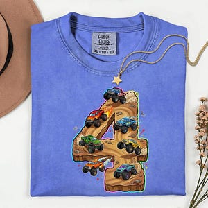 May include: A periwinkle blue Comfort Colors t-shirt with a colourful graphic of monster trucks racing on a brown number four. The shirt has a classic crew neck and short sleeves. A gold necklace with a star pendant is draped over the shirt.