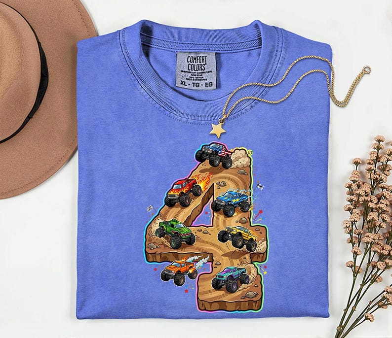 May include: A periwinkle blue Comfort Colors t-shirt with a colourful graphic of monster trucks racing on a brown number four. The shirt has a classic crew neck and short sleeves. A gold necklace with a star pendant is draped over the shirt.