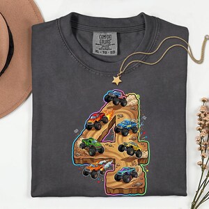 May include: Dark grey Comfort Colors t-shirt with a colourful graphic of monster trucks racing on a track shaped like the number 4. A gold star necklace and a brown hat are also in the image.