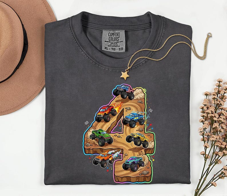 May include: Dark grey Comfort Colors t-shirt with a colourful graphic of monster trucks racing on a track shaped like the number 4. A gold star necklace and a brown hat are also in the image.