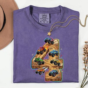 May include: A lilac t-shirt with a vibrant graphic of the number 4 and monster trucks. The design showcases monster trucks in red, green, blue, and yellow, navigating a brown terrain. A gold necklace with a star pendant is on the shirt.