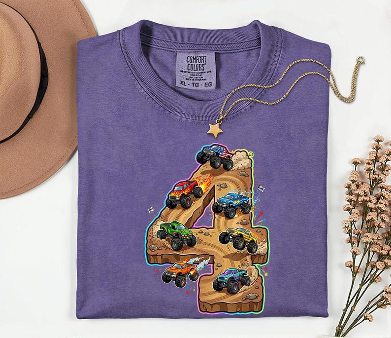 May include: A lilac t-shirt with a vibrant graphic of the number 4 and monster trucks. The design showcases monster trucks in red, green, blue, and yellow, navigating a brown terrain. A gold necklace with a star pendant is on the shirt.