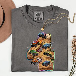 May include: Grey t-shirt featuring a colourful graphic of monster trucks racing on a track shaped like the number 4. The shirt has a crew neck and a tag that says "Comfort Colors". A gold necklace with a star pendant is draped on the shirt.