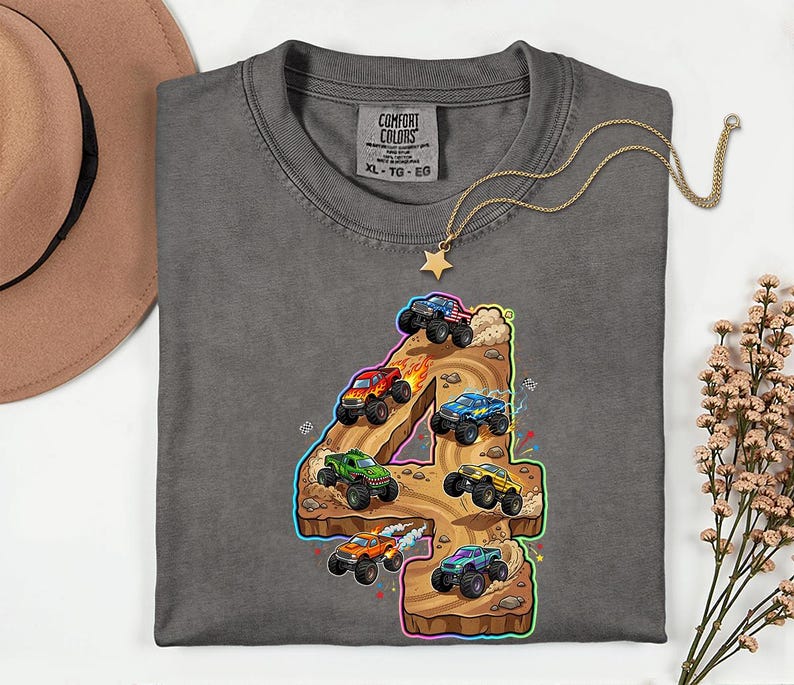 May include: Grey t-shirt featuring a colourful graphic of monster trucks racing on a track shaped like the number 4. The shirt has a crew neck and a tag that says "Comfort Colors". A gold necklace with a star pendant is draped on the shirt.