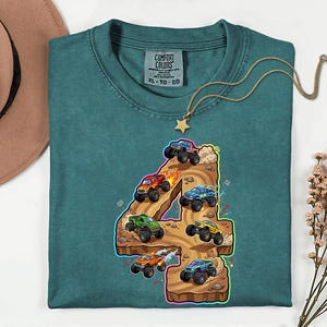 May include: Teal Comfort Colors t-shirt with a graphic of monster trucks racing on a number 4. The design includes monster trucks in red, blue, green, and orange. A gold necklace with a star pendant is draped on the shirt.