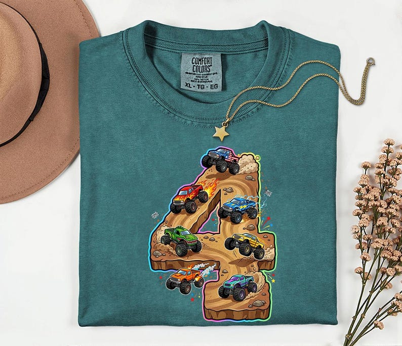 May include: Teal Comfort Colors t-shirt with a graphic of monster trucks racing on a number 4. The design includes monster trucks in red, blue, green, and orange. A gold necklace with a star pendant is draped on the shirt.