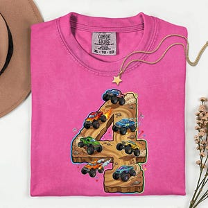 May include: A vibrant pink Comfort Colors t-shirt with a graphic of monster trucks forming the number 4. The trucks are in different colours, driving on a brown terrain. A gold necklace with a star pendant is draped across the shirt.