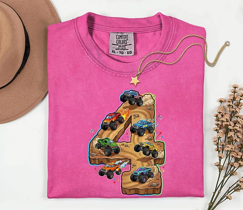 May include: A vibrant pink Comfort Colors t-shirt with a graphic of monster trucks forming the number 4. The trucks are in different colours, driving on a brown terrain. A gold necklace with a star pendant is draped across the shirt.