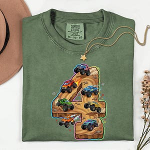 May include: Green t-shirt featuring a vibrant graphic of monster trucks racing around the number 4. The shirt has a crew neck and short sleeves. The Comfort Colors label is visible at the neckline.