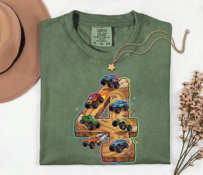 May include: Green t-shirt featuring a vibrant graphic of monster trucks racing around the number 4. The shirt has a crew neck and short sleeves. The Comfort Colors label is visible at the neckline.
